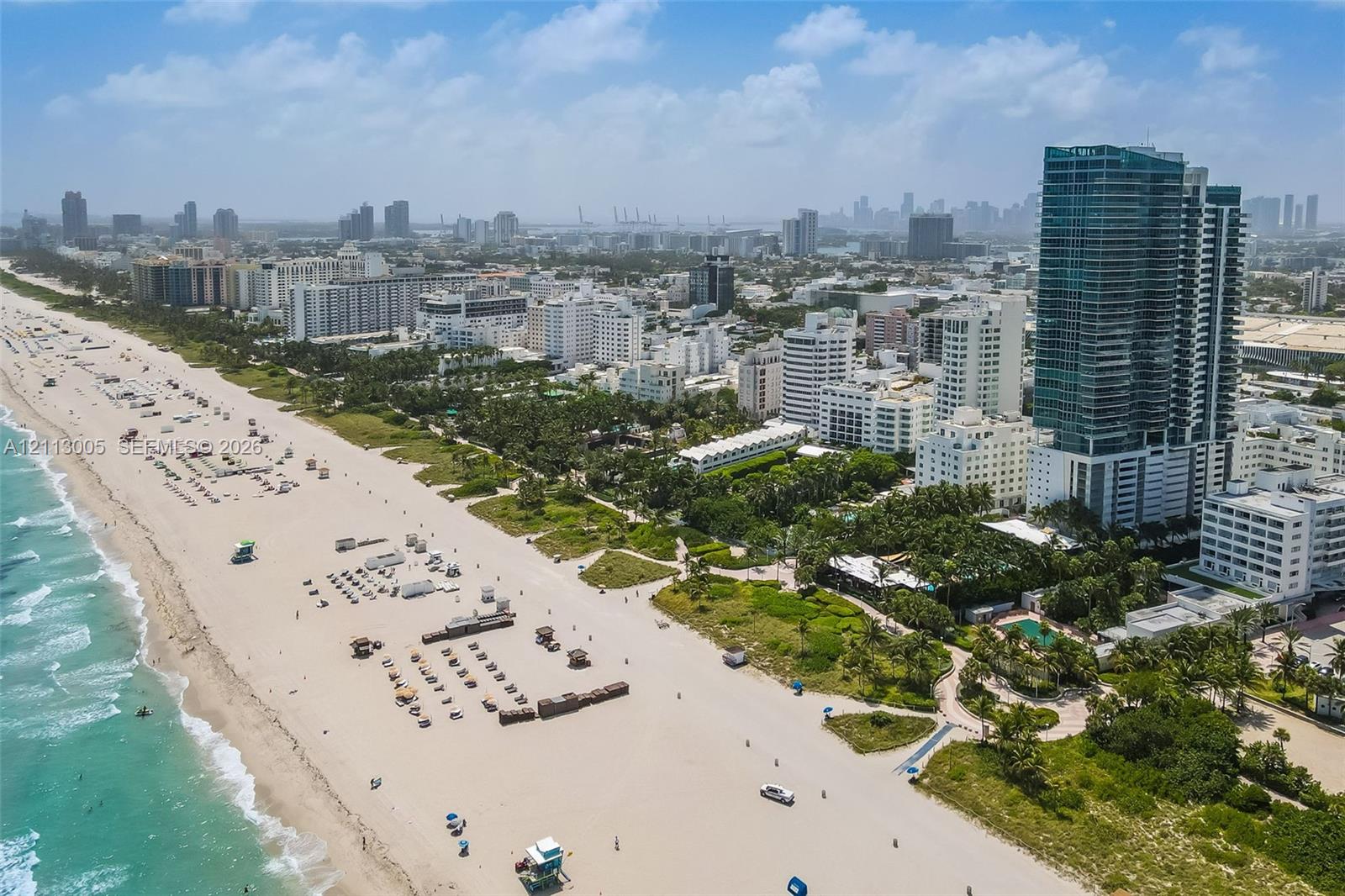 Aerial perspective featuring immediate oceanfront positioning, expansive sandy shoreline, dense coastal foliage, and high-rise urban density