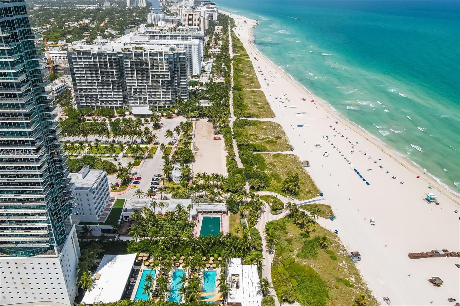 Aerial perspective of a high-rise residential building featuring multiple ground-level swimming pools, manicured palm landscaping, and direct access to the coastline
