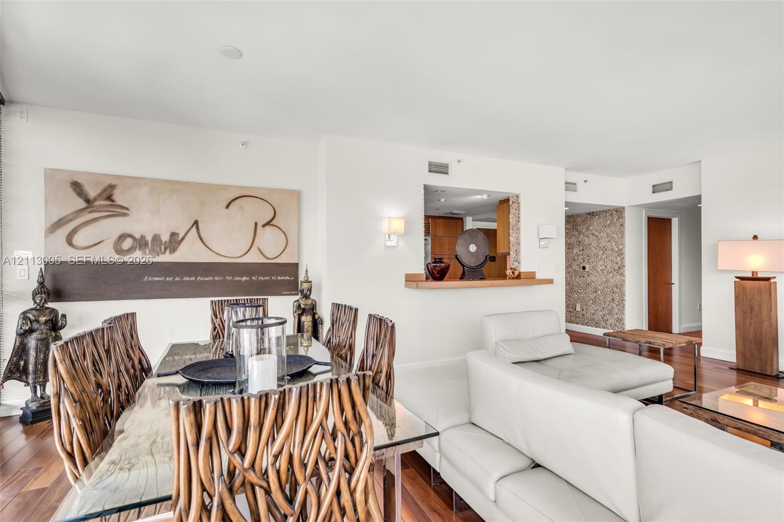 Open-concept living space featuring wood-finish flooring, a pass-through kitchen window with a built-in wooden ledge, wall-mounted light fixtures, and a decorative textured accent wall