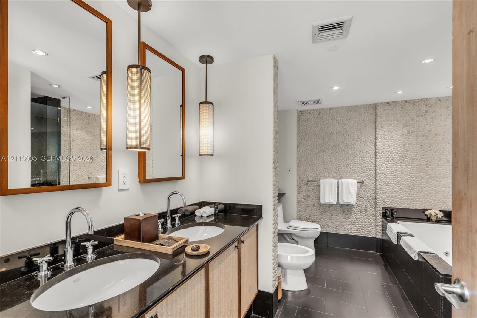 Dual-sink vanity with dark stone countertop, textured stone wall accents, recessed lighting, and modern cylindrical pendant fixtures