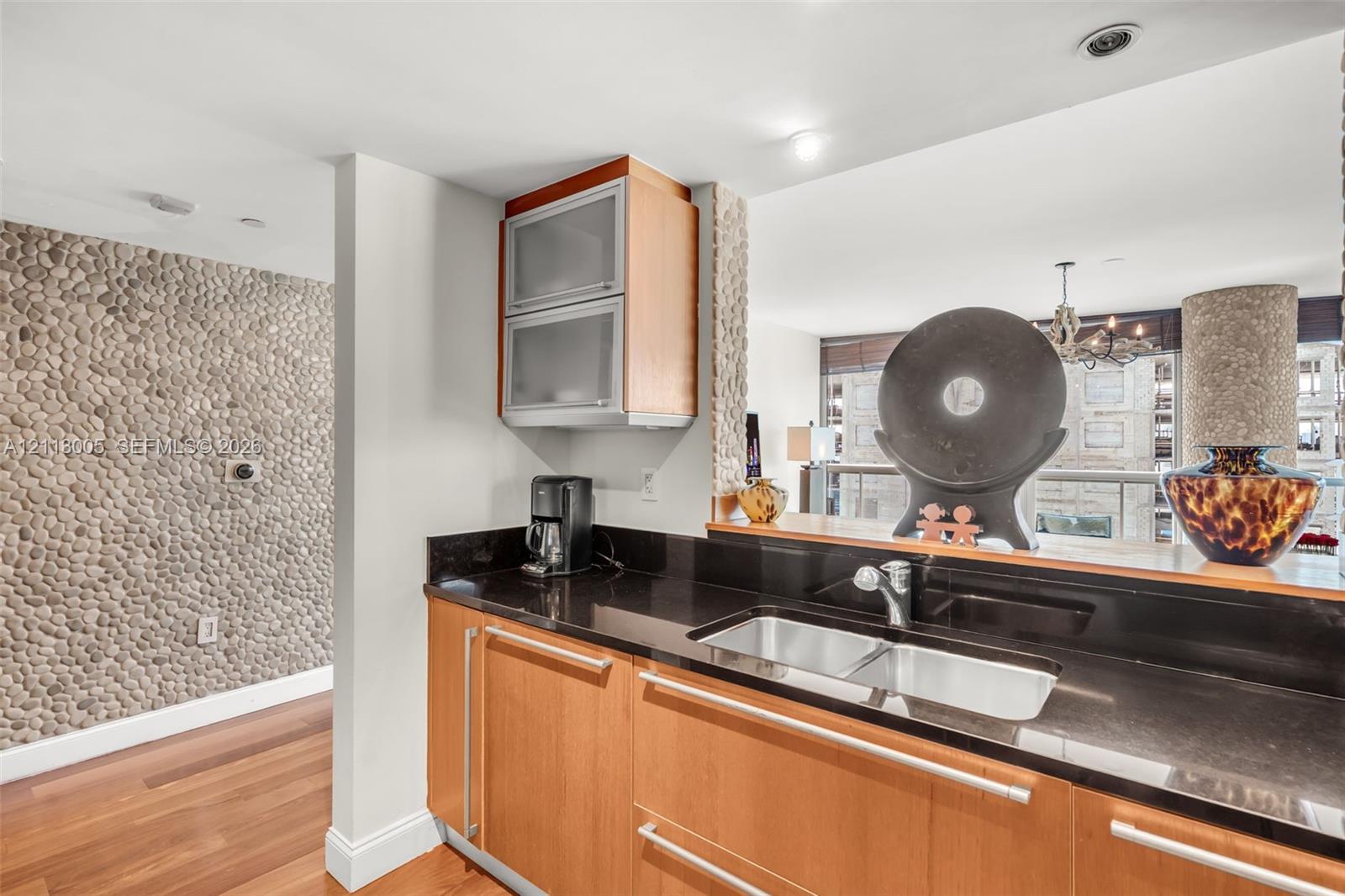 Kitchen area featuring wood cabinetry, dark stone countertops, an integrated double-basin stainless steel sink, frosted glass upper cabinets, and a decorative pebble-tile accent wall