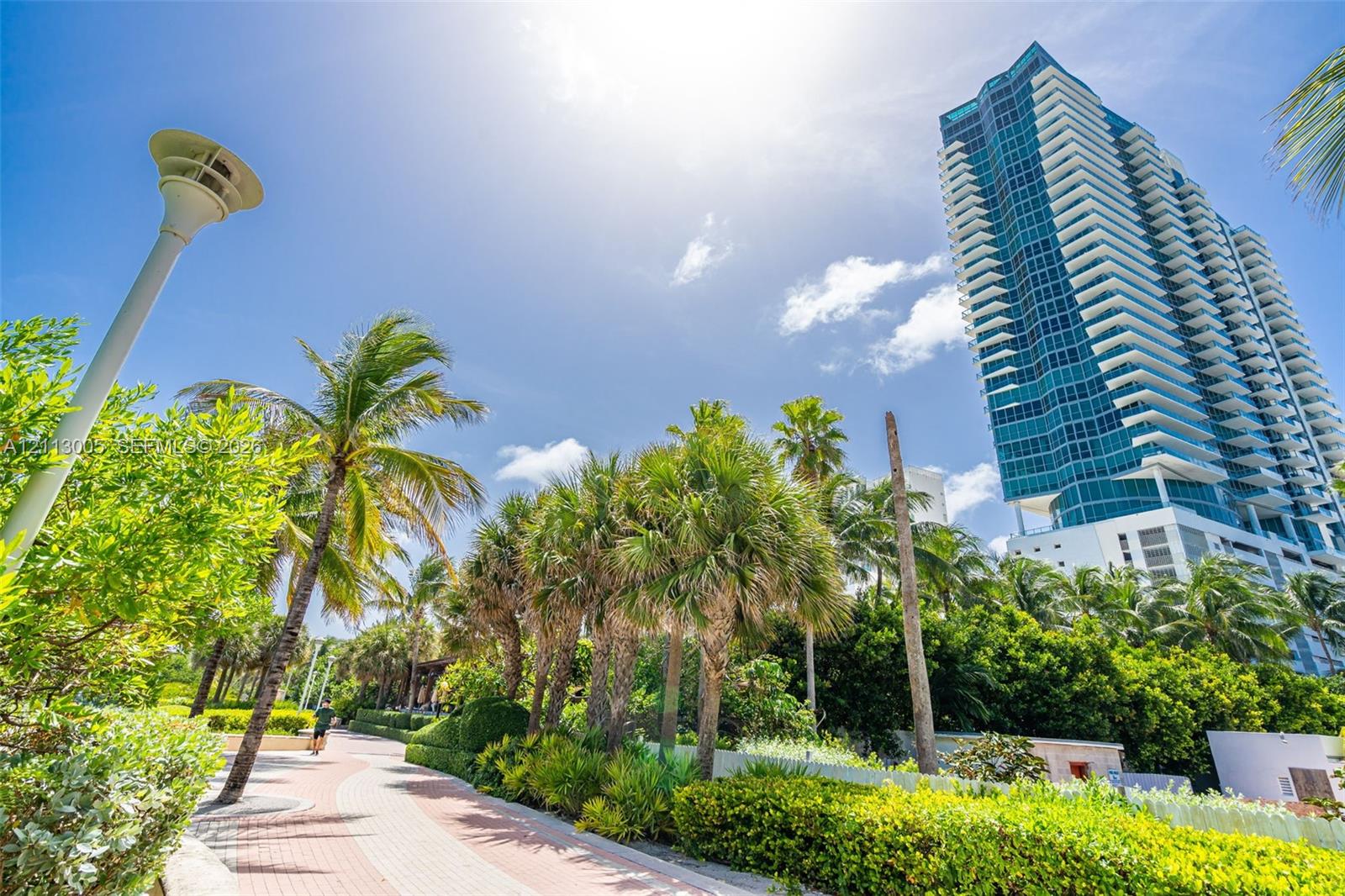 High-rise residential tower featuring blue glass cladding and tiered balconies, situated along a landscaped pedestrian walkway with mature palm trees and manicured shrubbery