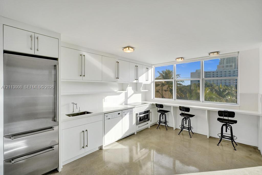 Kitchen featuring white cabinetry with integrated hardware, polished concrete flooring, stainless steel refrigerator, built-in dishwasher, and a window-lined counter