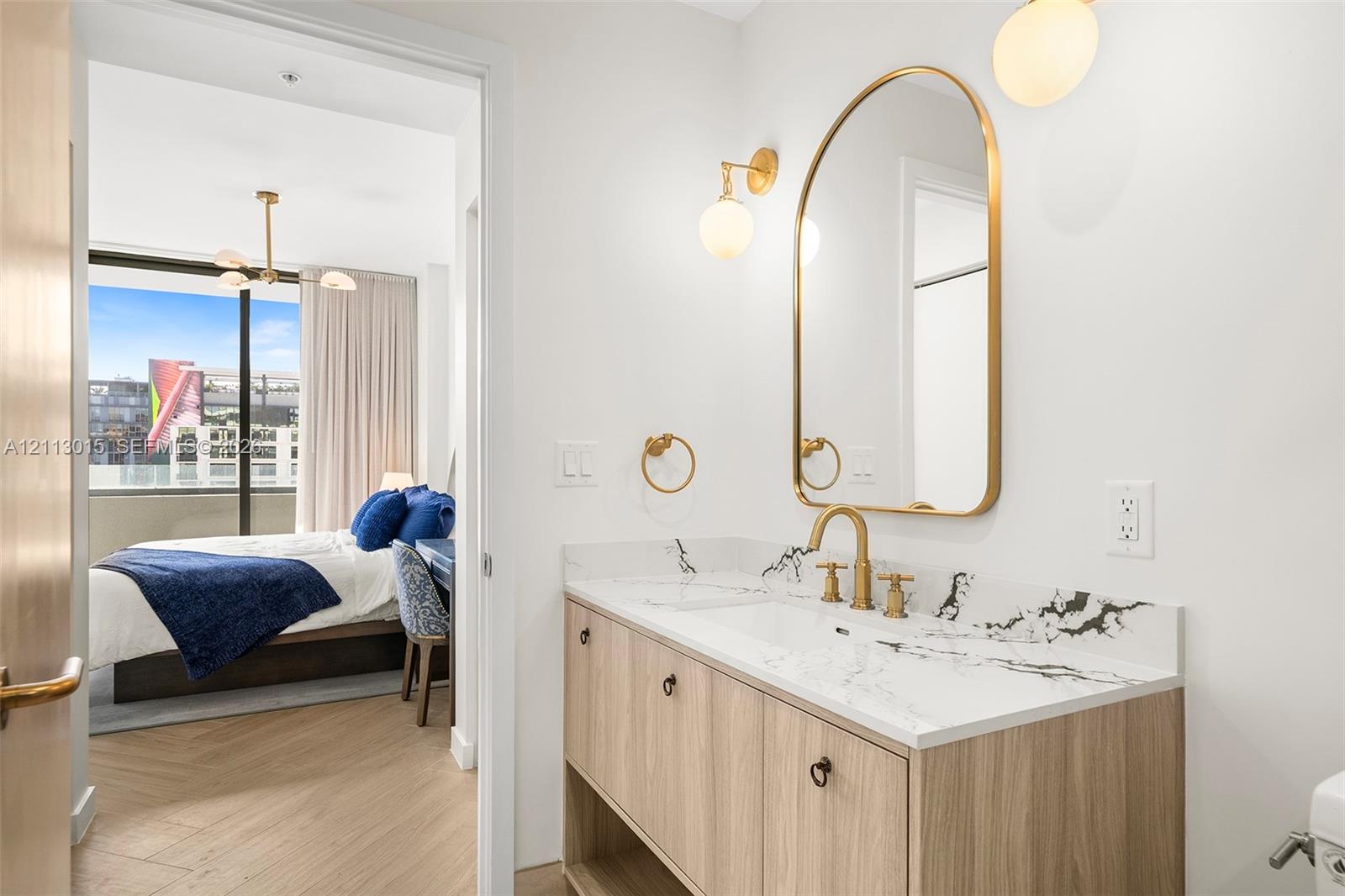 Modern vanity with a veined stone countertop and integrated sink, complemented by a brass-finished arched mirror, wall-mounted globe lighting, and light wood-finish flooring
