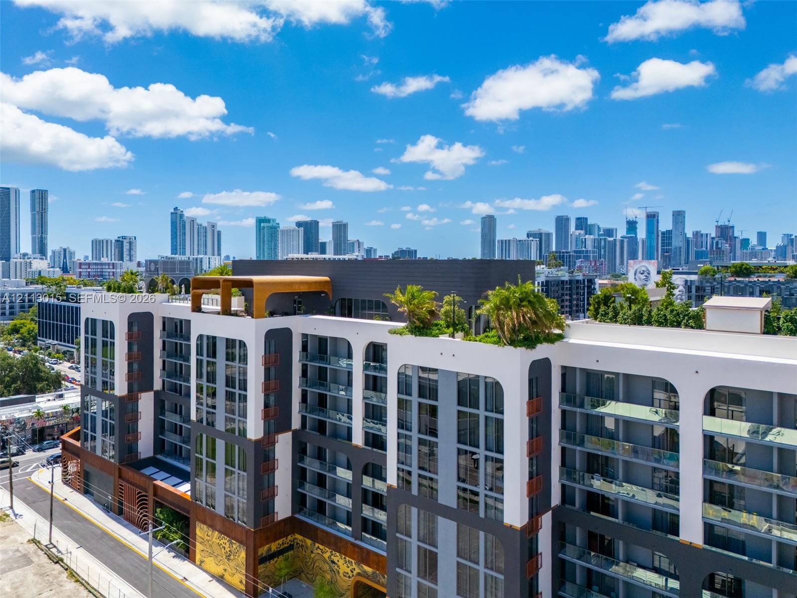 Modern multi-story building facade featuring arched architectural details, private glass-railed balconies, a rooftop terrace with mature landscaping, and multi-toned exterior paneling