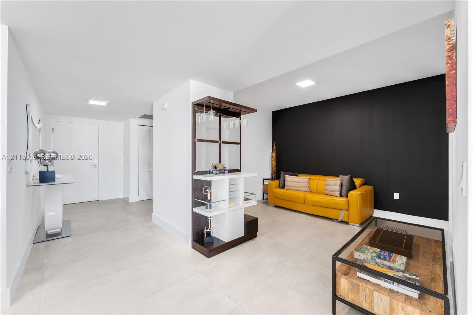 Interior space featuring tile flooring, recessed ceiling lighting, a high-contrast black accent wall, and a custom built-in wood and white laminate bar cabinet with glass shelving Interior space featuring tile flooring, recessed ceiling lighting, a high-contrast black accent wall, and a custom built-in wood and white laminate bar cabinet with glass shelving