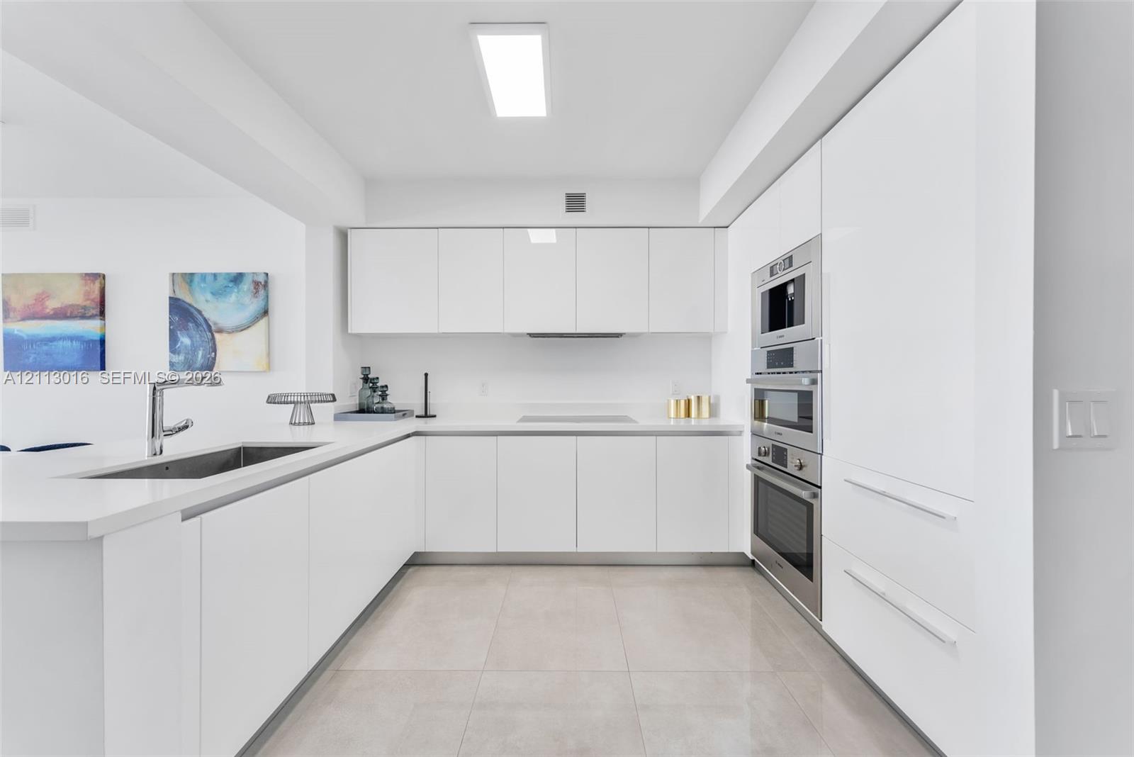 Minimalist kitchen featuring white flat-panel cabinetry, light-toned stone-finish countertops, stainless steel wall-mounted oven and microwave, and large-format tile flooring Minimalist kitchen featuring white flat-panel cabinetry, light-toned stone-finish countertops, stainless steel wall-mounted oven and microwave, and large-format tile flooring