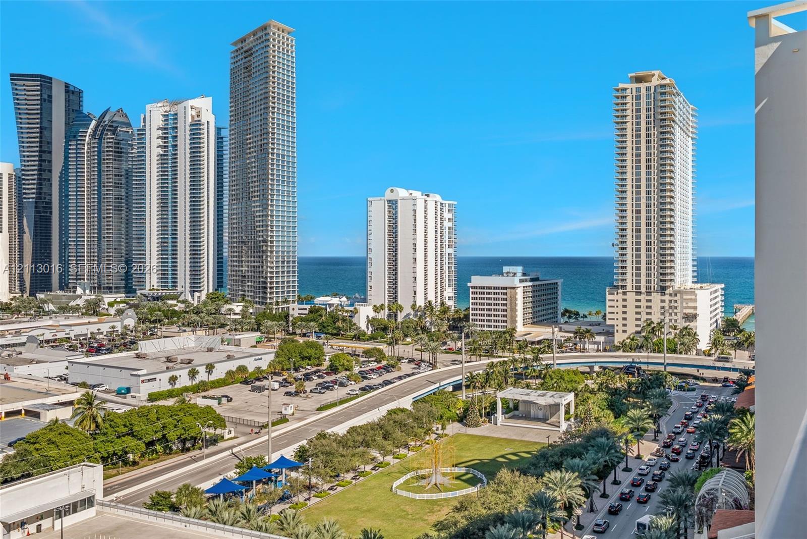 High-rise urban landscape featuring ocean water, multi-story glass-facade towers, manicured green space, and palm tree vegetation