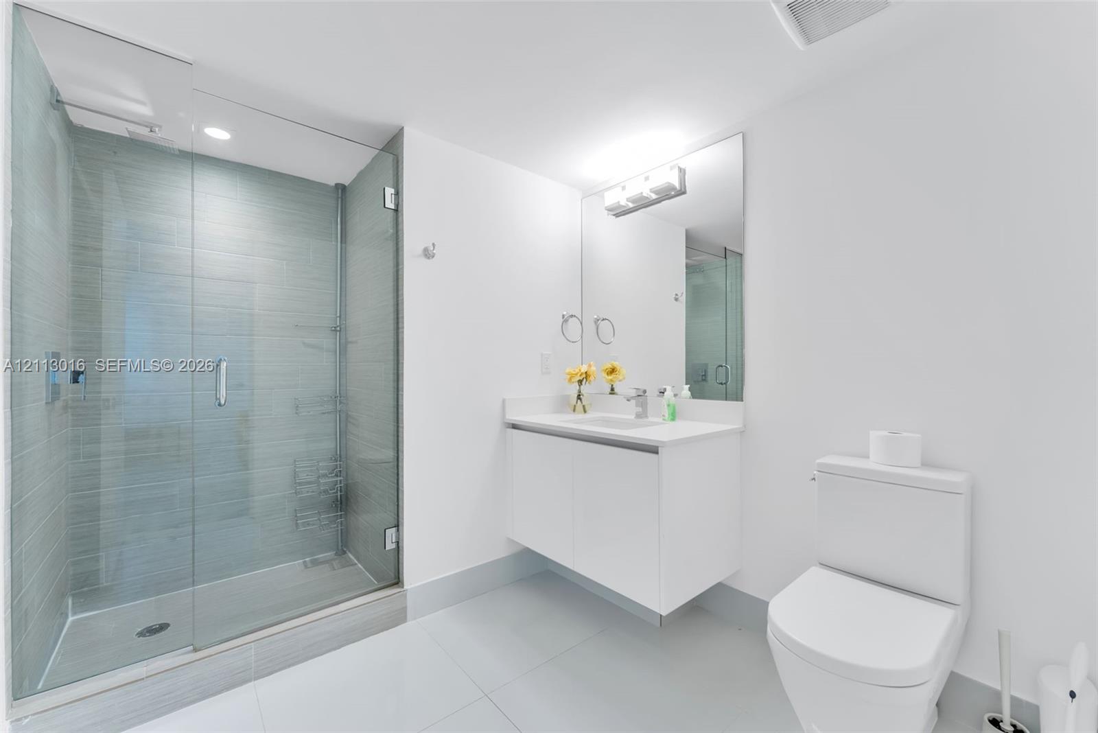Glass-enclosed walk-in shower with horizontal tile surround, wall-mounted white vanity, polished countertop with integrated sink, and vanity-width mirror with overhead lighting Glass-enclosed walk-in shower with horizontal tile surround, wall-mounted white vanity, polished countertop with integrated sink, and vanity-width mirror with overhead lighting