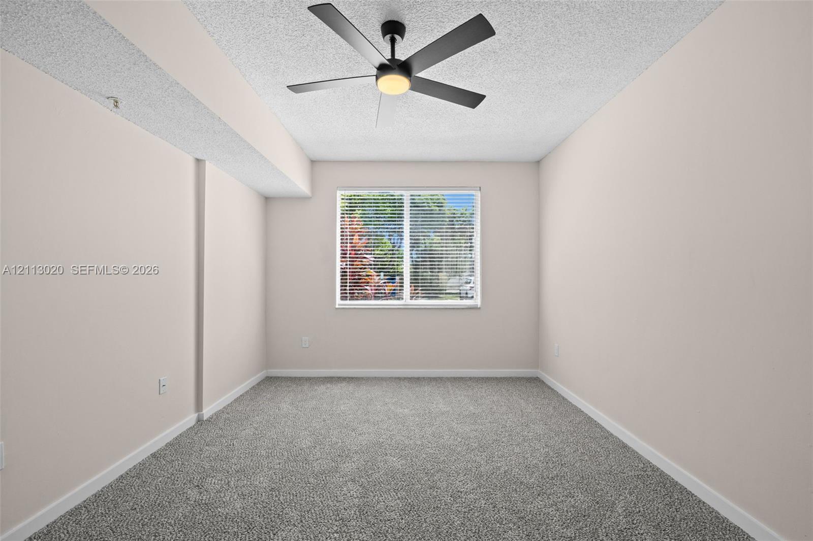 Neutral-toned interior featuring wall-to-wall carpeting, a multi-blade ceiling fan with integrated lighting, and a single-pane window with horizontal blinds