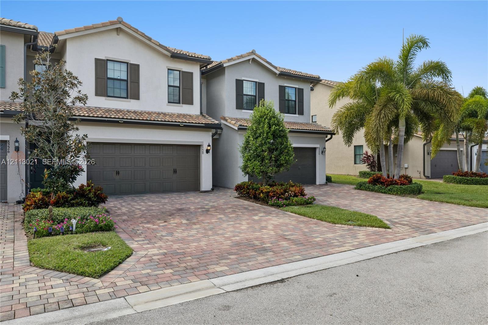 Two-story stucco exterior featuring a multi-gabled tile roof, paver driveway, attached two-car garage, and landscaped garden beds