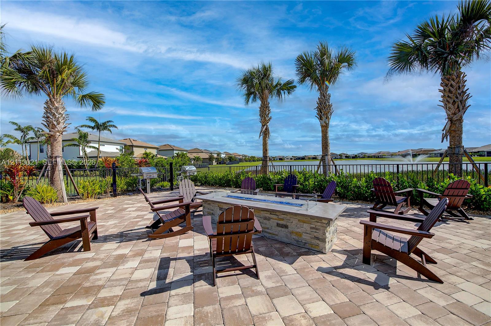 Paver patio featuring a stone-clad linear fire pit, metal outdoor grill, water-facing fence line, and mature palm trees