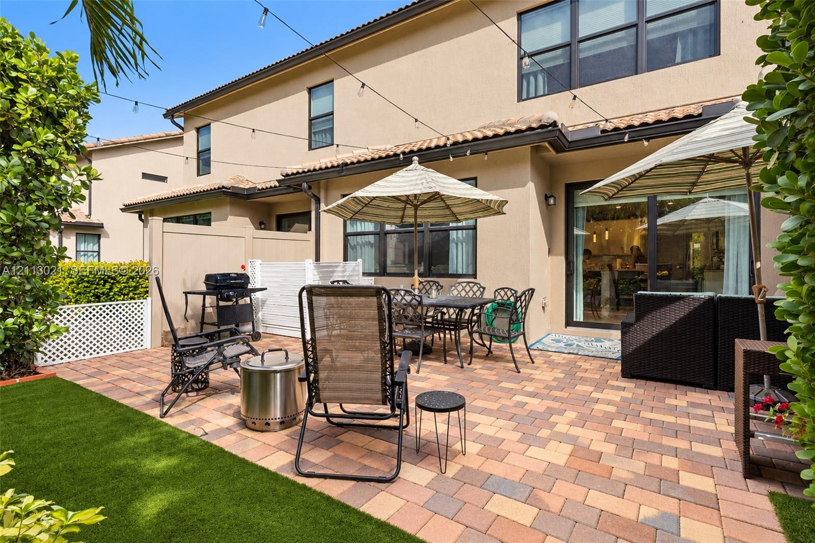 Rear exterior space featuring a paved stone patio, artificial turf lawn, privacy fencing with lattice detailing, and overhead string lighting