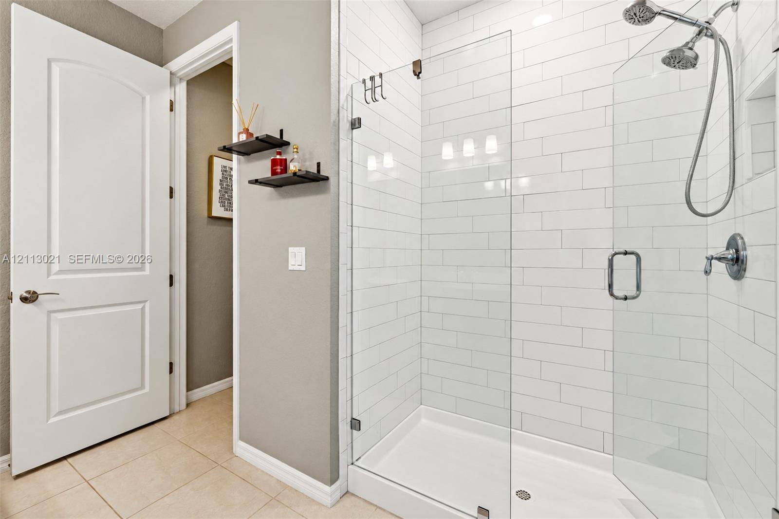 Walk-in shower with white subway tile surround, frameless glass door, dual shower heads, and built-in recessed shelf, complemented by large-format tile flooring and a solid-core interior door