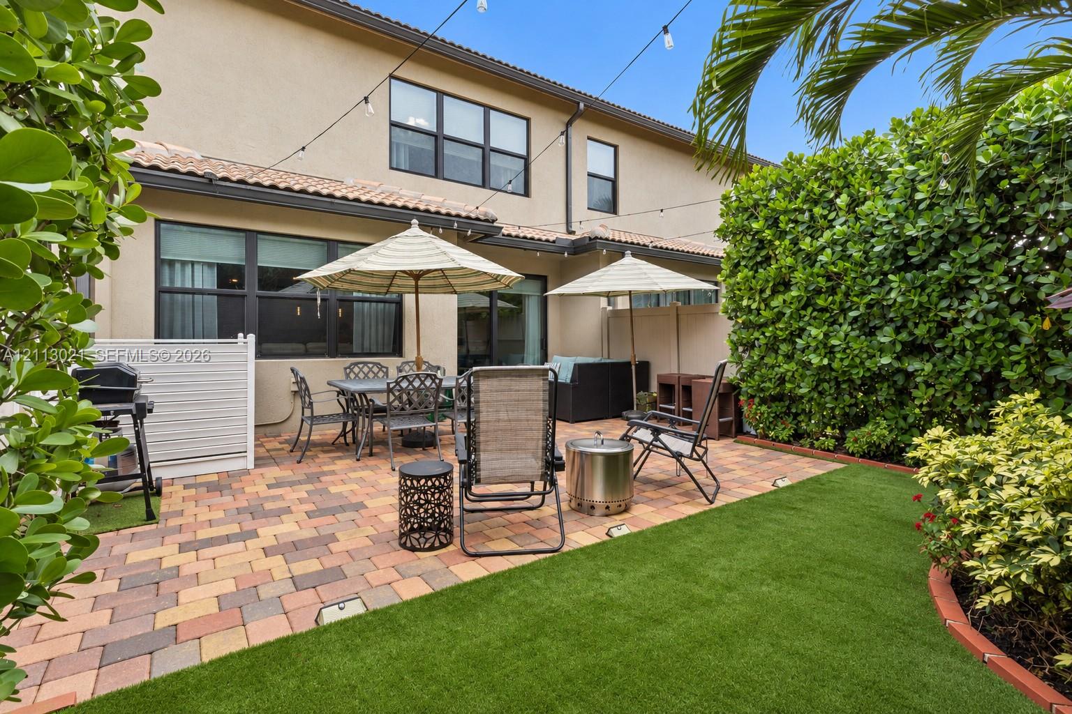 Rear patio featuring multicolored paver hardscaping, synthetic turf lawn, privacy hedging, and overhead string lighting