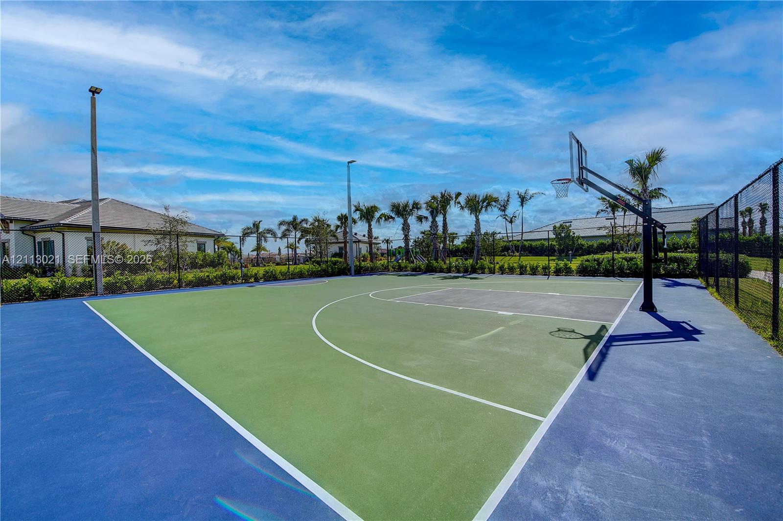Outdoor basketball court featuring a multi-tone hard surface, professional hoop system, perimeter fencing, and integrated pole lighting