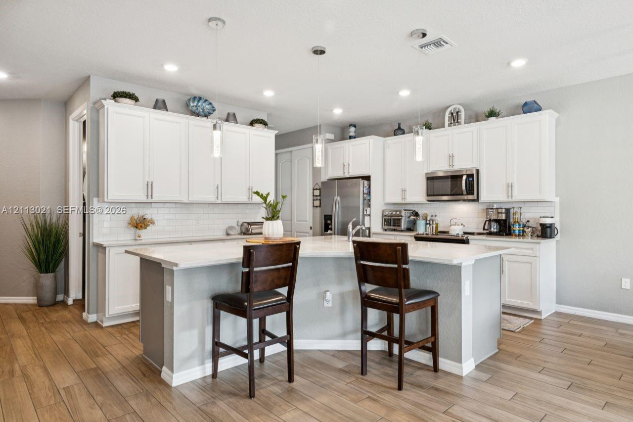 Kitchen featuring white shaker-style cabinetry, a central island with a solid-surface countertop, white subway tile backsplash, stainless steel appliances, and wood-finish flooring Kitchen featuring white shaker-style cabinetry, a central island with a solid-surface countertop, white subway tile backsplash, stainless steel appliances, and wood-finish flooring