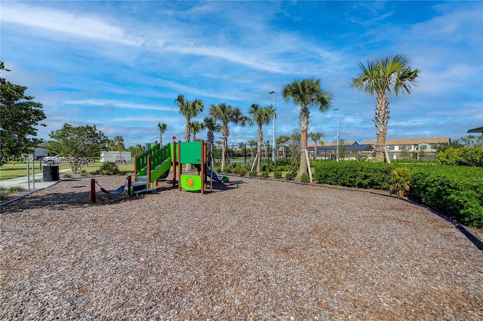 Outdoor recreational area featuring a structured playground, wood chip surfacing, manicured perimeter shrubbery, and mature palm trees