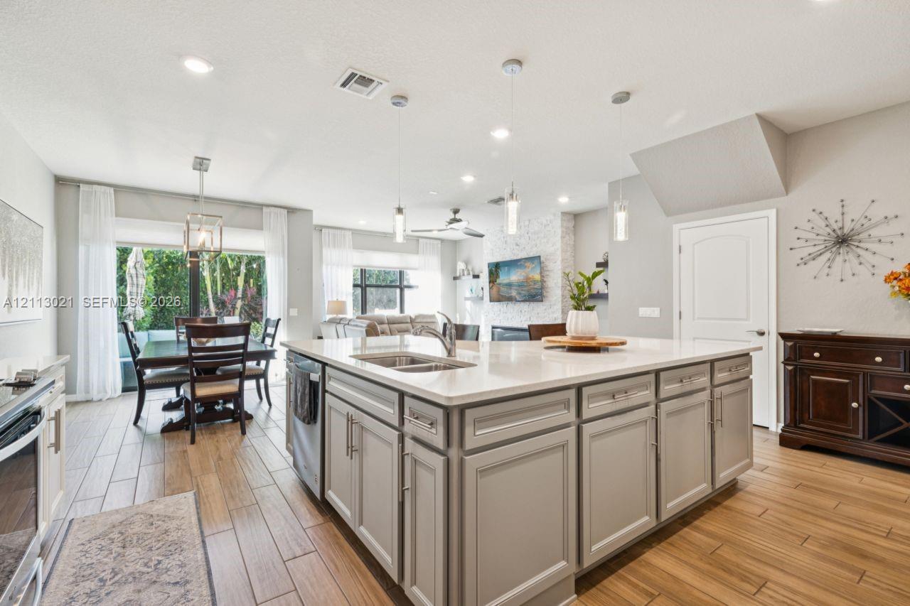 Kitchen featuring a large central island with light-toned cabinetry, solid surface countertops, a stainless steel double-basin undermount sink, pendant lighting, and wood-finish flooring Kitchen featuring a large central island with light-toned cabinetry, solid surface countertops, a stainless steel double-basin undermount sink, pendant lighting, and wood-finish flooring