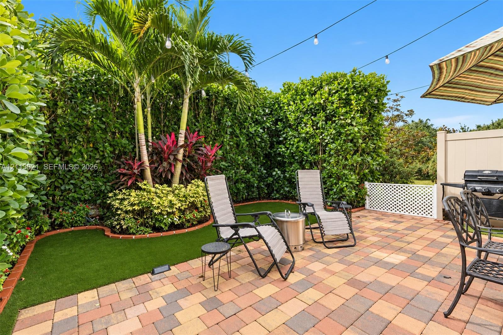 Backyard featuring multicolor paver patio, synthetic turf, mature palm trees, perimeter privacy hedging, and overhead string lighting