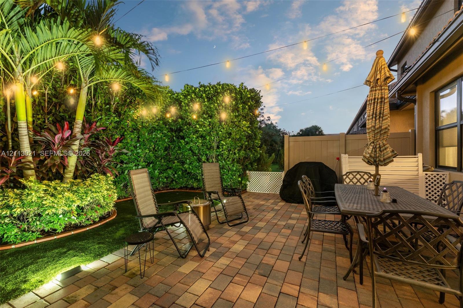 Rear patio featuring multi-tone pavers, mature tropical landscaping, artificial turf, perimeter fencing, and overhead string lighting