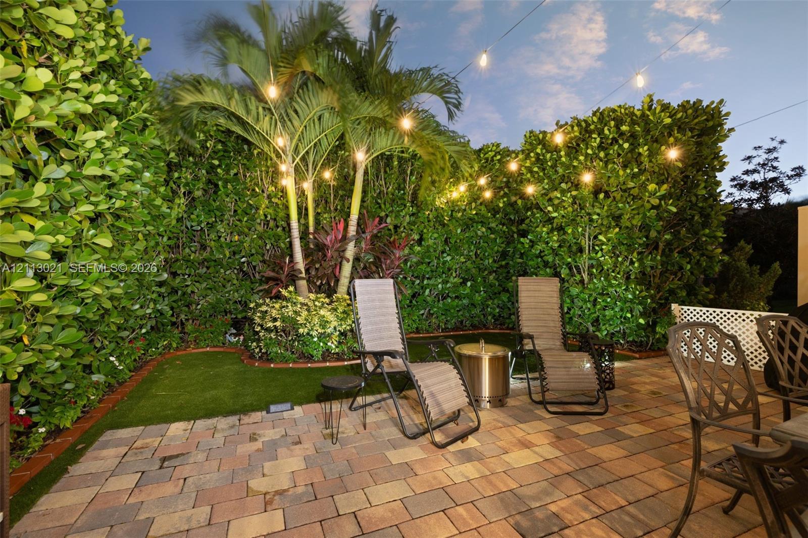 Private outdoor space featuring a paved brick patio, manicured synthetic lawn, perimeter privacy hedges, and palm trees
