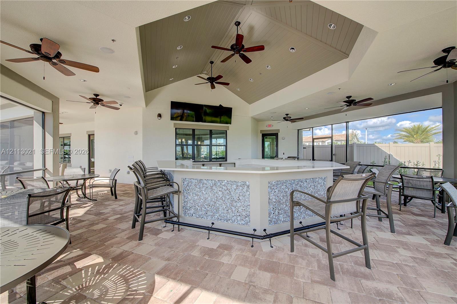 Covered outdoor patio featuring a vaulted ceiling with tongue-and-groove detailing, custom built-in bar with stone-tiled facade, paver flooring, and multiple ceiling fans