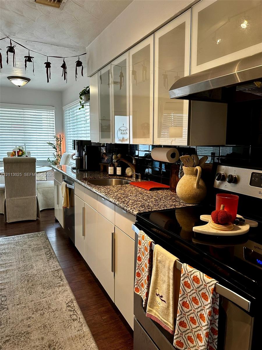 Kitchen featuring flat-panel cabinetry with glass-front upper doors, stone-finish countertops, black tile backsplash, stainless steel range with overhead vent hood, and wood-finish flooring