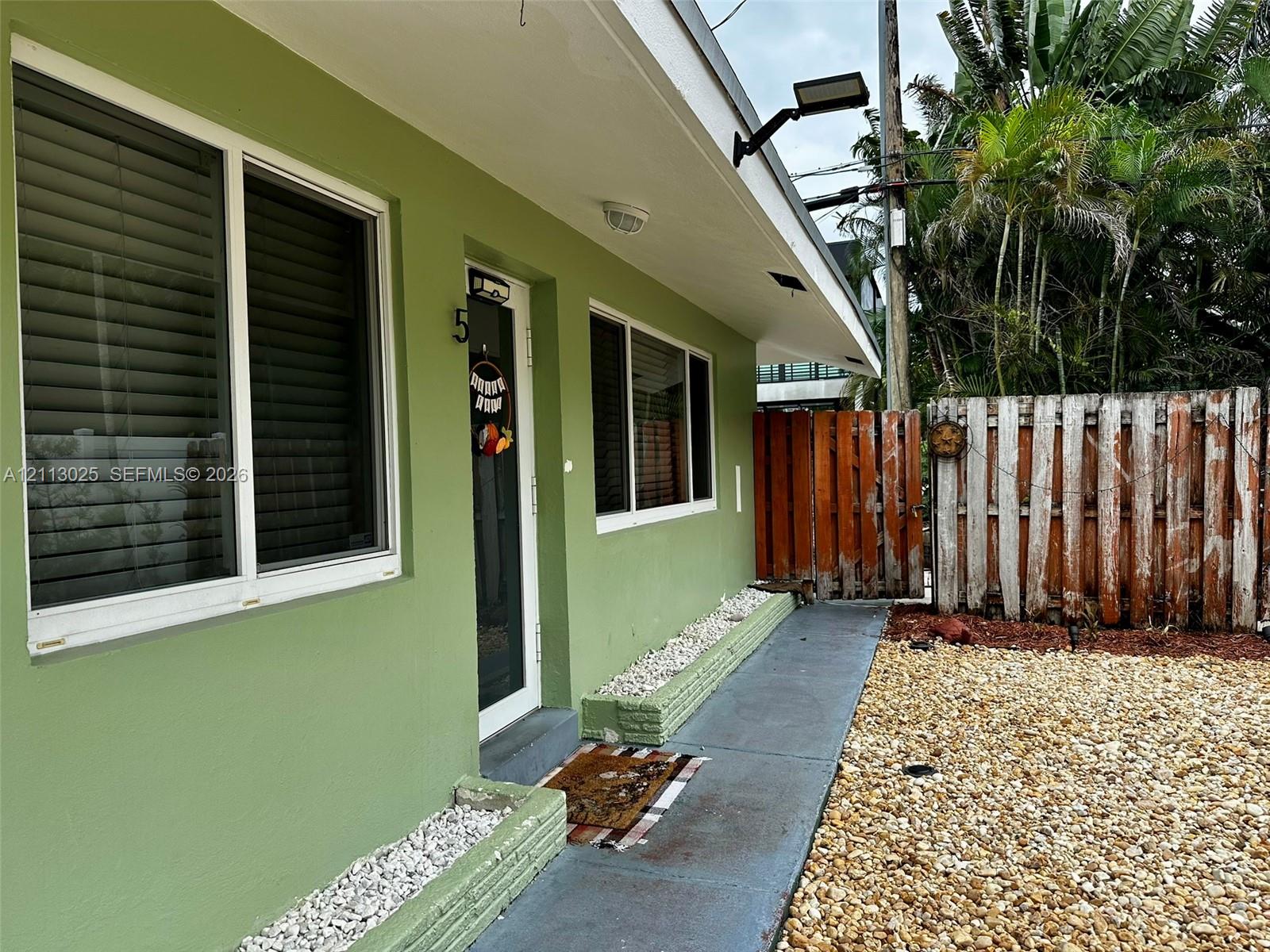 Exterior facade featuring smooth-finish stucco, horizontal sliding windows with integrated blinds, a dedicated entry door, decorative stone-filled masonry planters, and a fenced yard with graveled landscaping