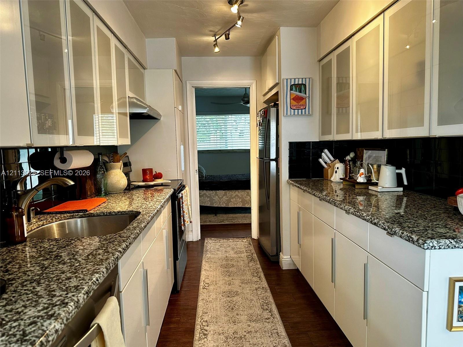 Galley kitchen featuring stone countertops, glass-panel upper cabinetry, stainless steel appliances, dark tile backsplash, and track lighting