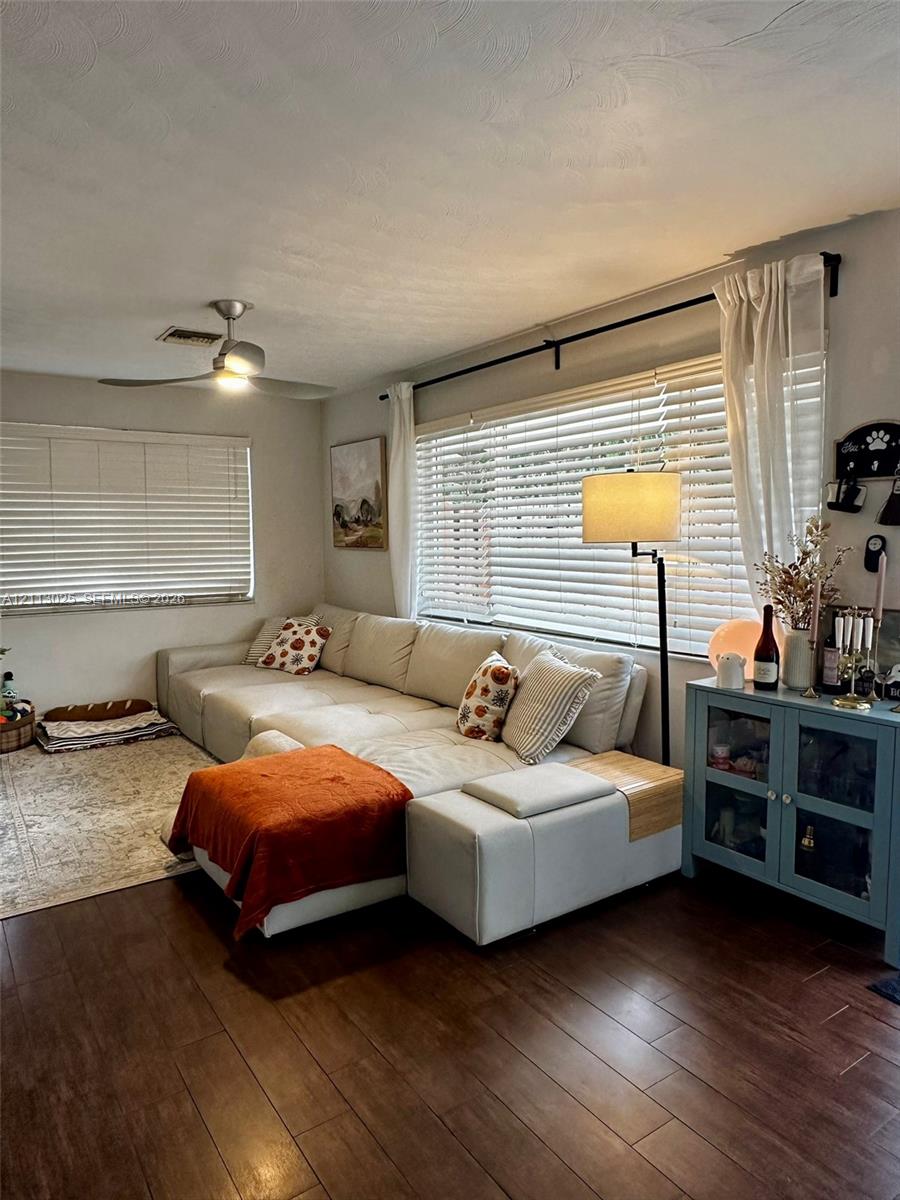 Living area featuring dark wood-finish flooring, two large windows with horizontal blinds, and a brushed nickel ceiling fan with integrated lighting
