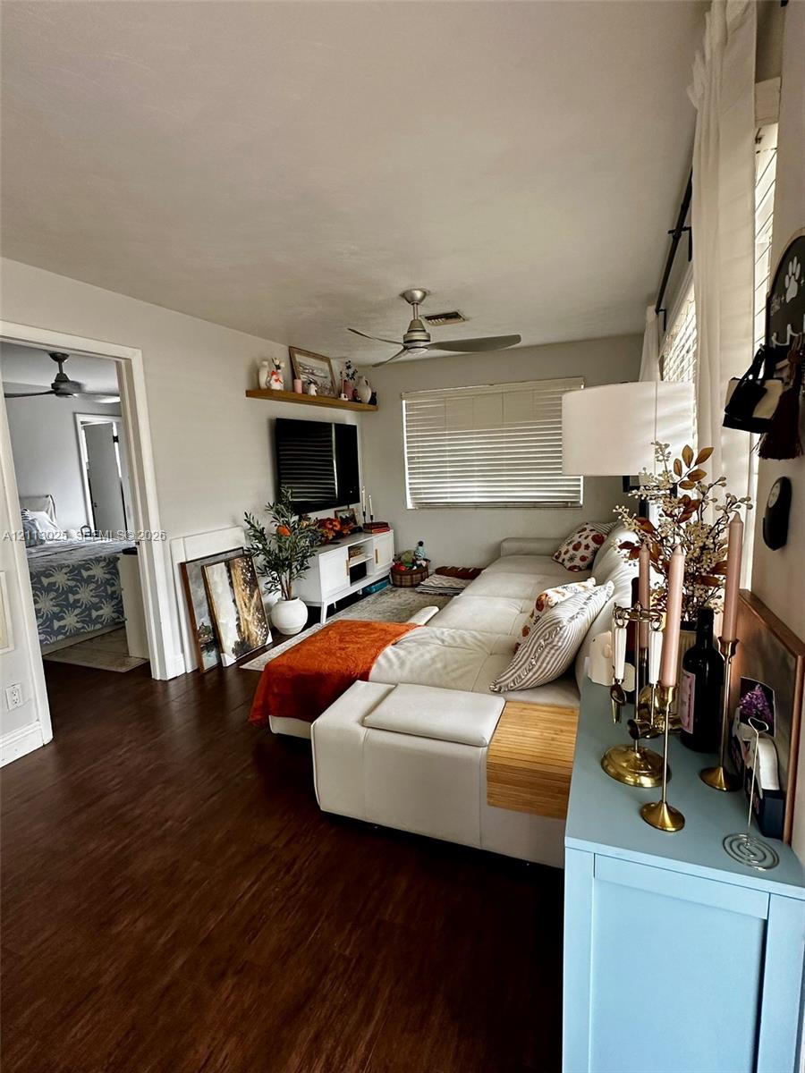 Living space featuring dark wood-finish flooring, a wall-mounted floating shelf, a ceiling fan, and horizontal window blinds