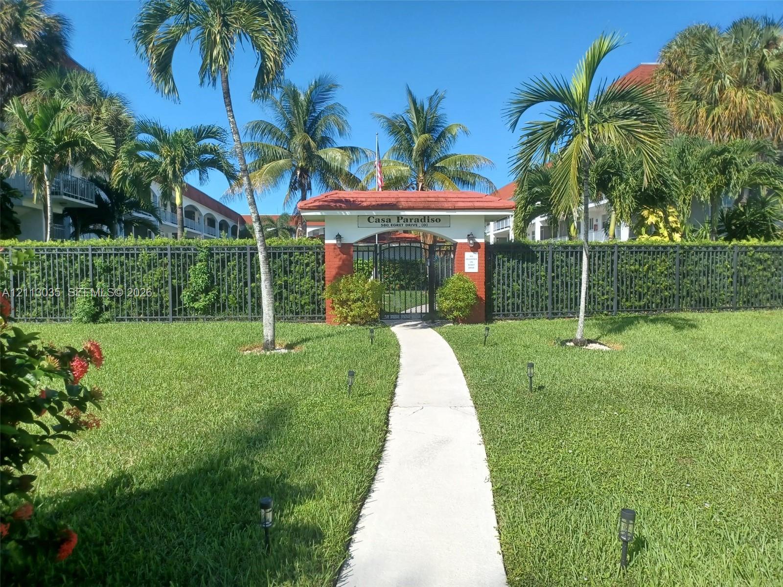 Gated entrance featuring a tile-roofed masonry archway, concrete pedestrian path, integrated landscape lighting, perimeter metal fencing, and mature palm trees