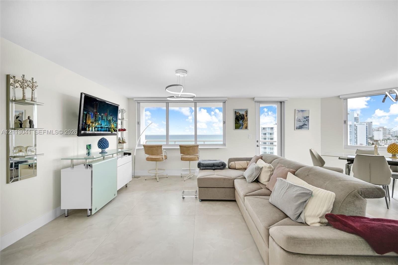 Open-concept living area featuring large-format tile flooring, a modern sculptural chandelier, floor-to-ceiling windows with coastal scenery, and a glass-paneled entry door