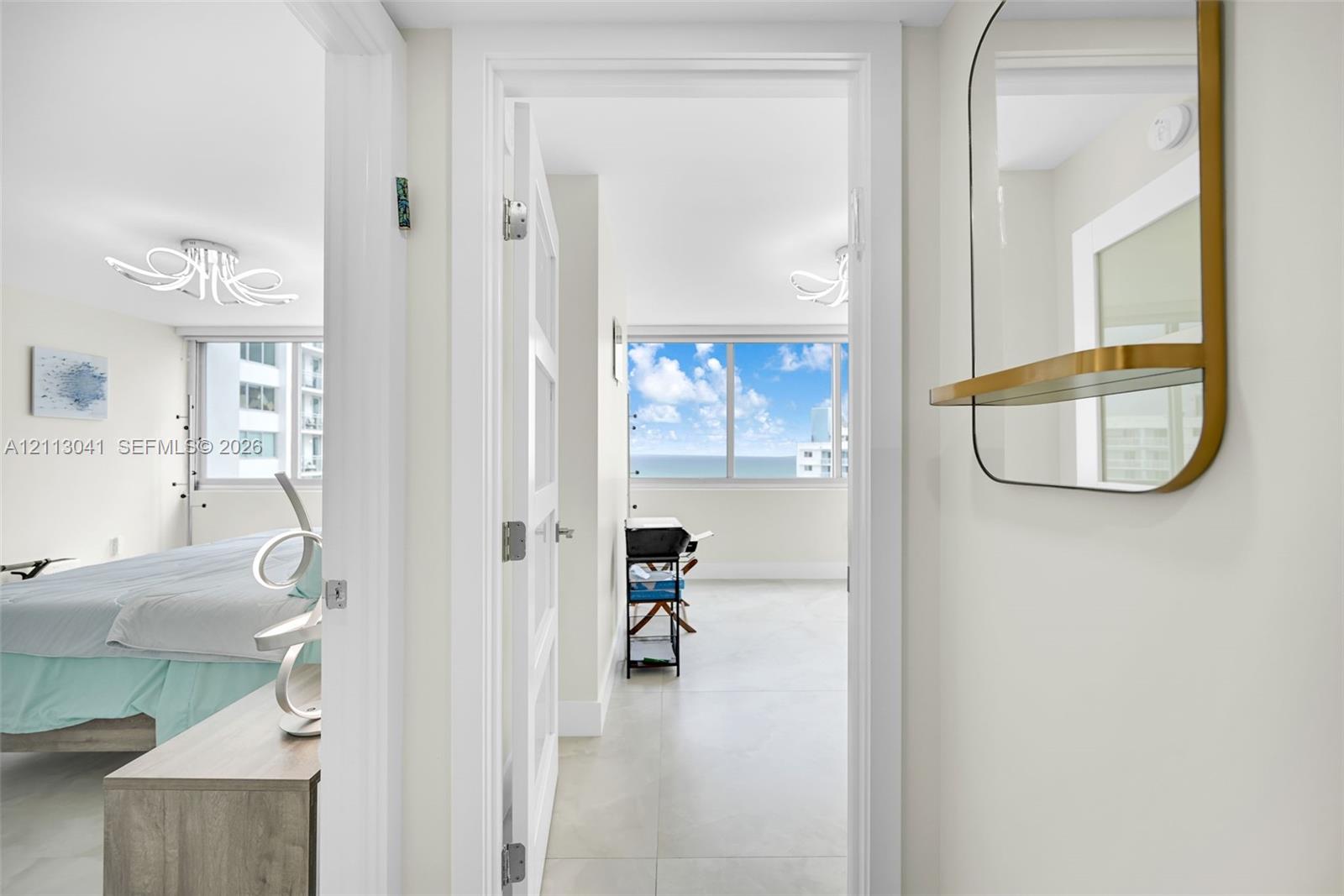 Light-toned large-format tile flooring, arched decorative ceiling lighting fixtures, a gold-framed wall-mounted mirror with an integrated shelf, and floor-to-ceiling windows with water scenery