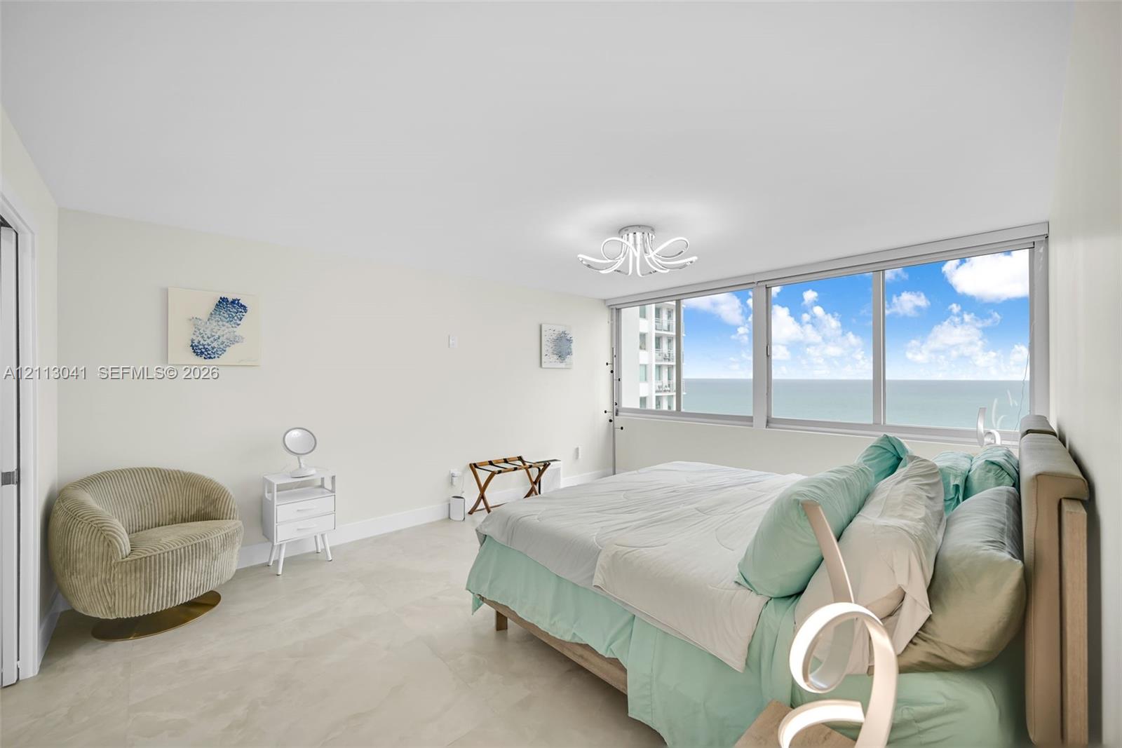 Expansive bedroom featuring large-format tile flooring, floor-to-ceiling windows with ocean and coastal scenery, and a contemporary multi-arm flush-mount light fixture