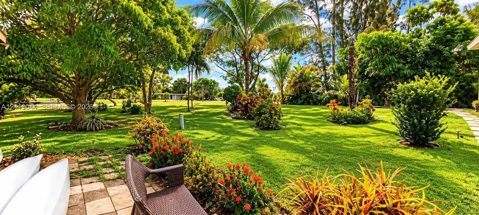 Expansive backyard featuring mature shade trees, established ornamental landscaping, palm trees, and a paved stone patio