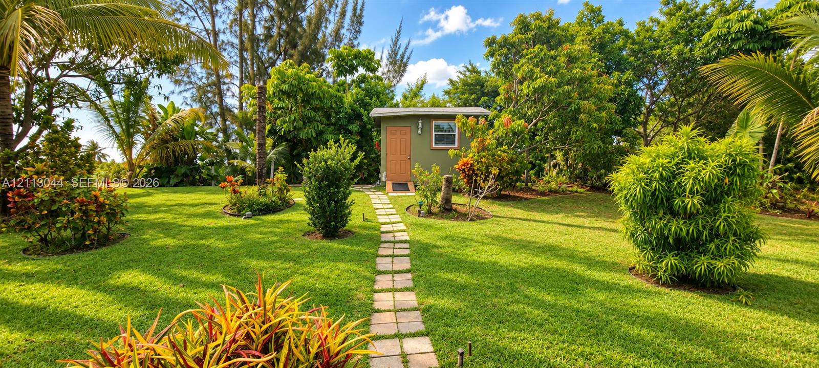 Expansive lawn featuring mature tropical landscaping, a stone-paver walkway, and a detached exterior structure with a central entry door and window