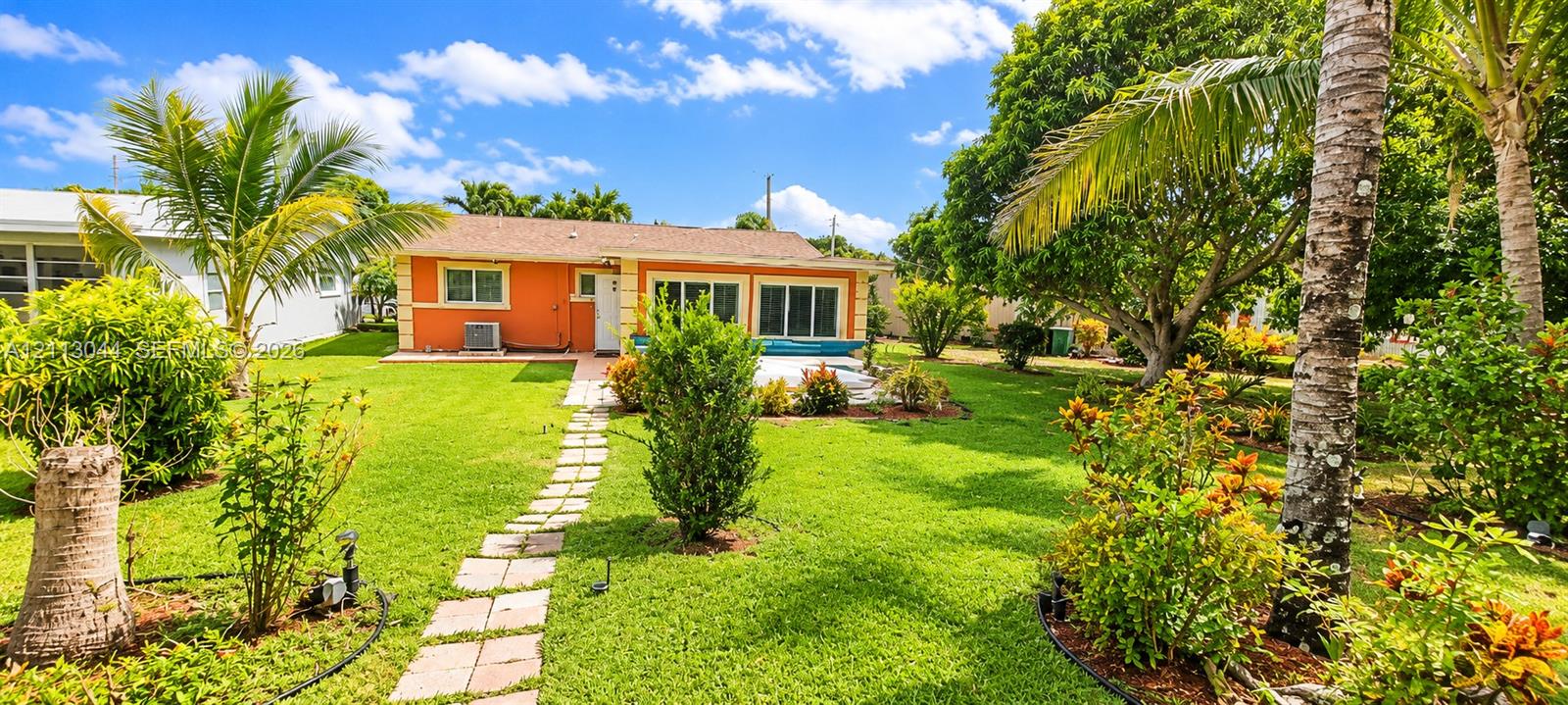 Single-story residence featuring a stucco exterior, shingled roof, stone-paver walkway, expansive lawn, and established tropical landscaping