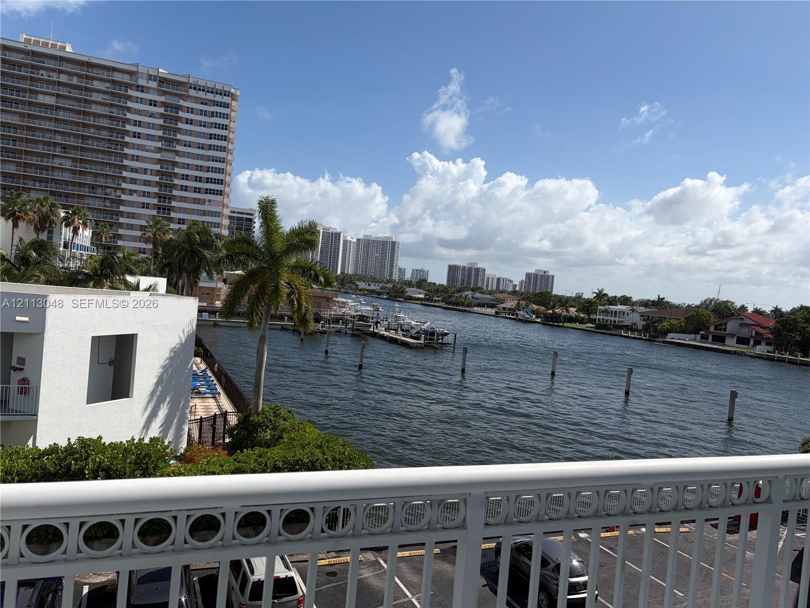 Exterior balcony featuring a decorative metal railing overlooking a waterway, mature palm trees, and coastal urban scenery