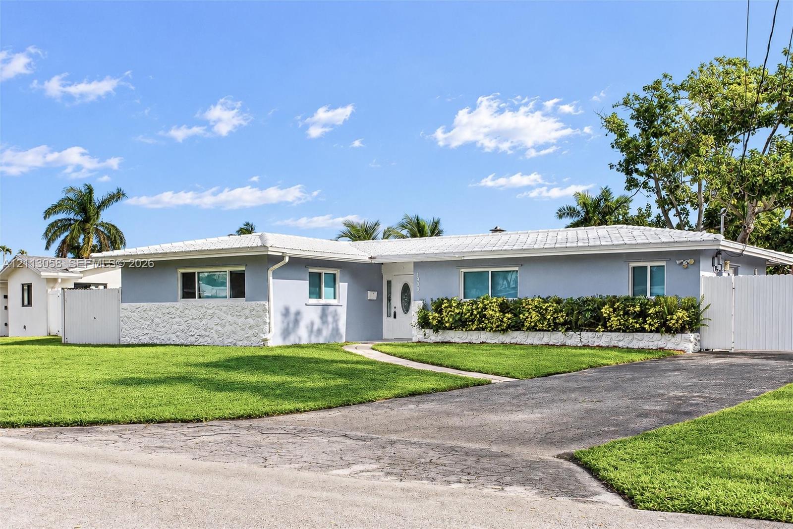 Single-story exterior featuring a light-colored stucco facade with stone-textured skirting, a white-tiled roof, an asphalt driveway, and a white privacy fence