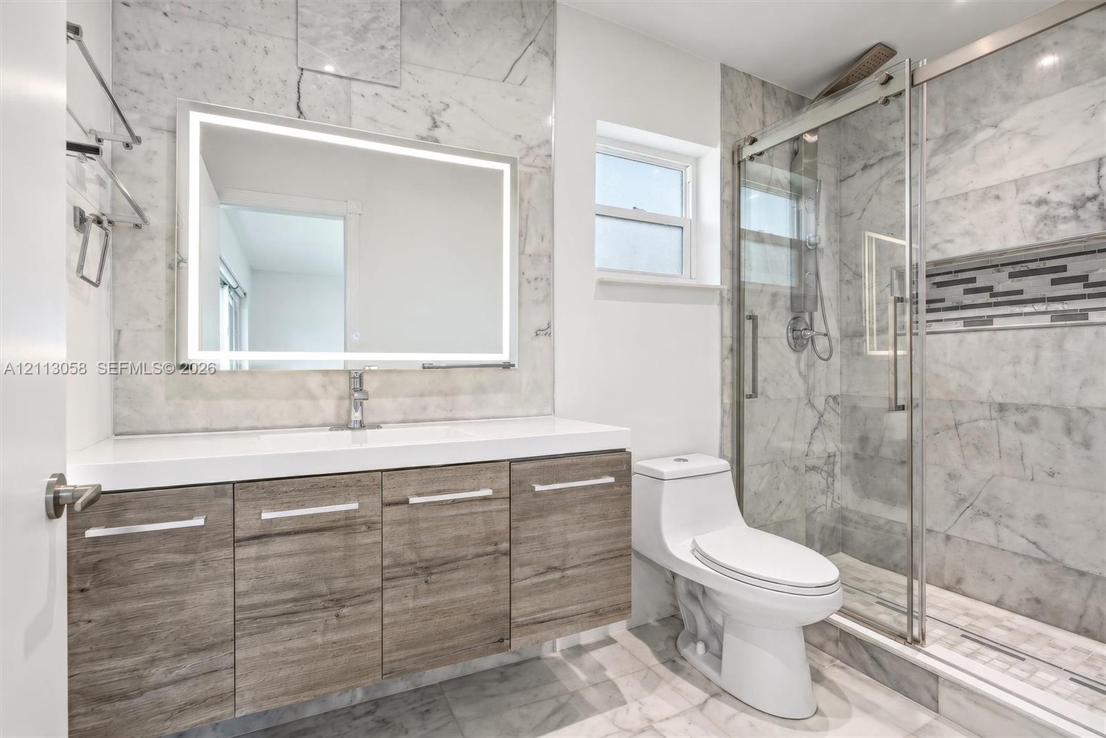 Modern bathroom featuring wood-finish vanity cabinetry, a white countertop with an integrated sink, floor-to-ceiling stone-tiled walls, a backlit mirror, and a glass-enclosed shower with a decorative mosaic tile accent