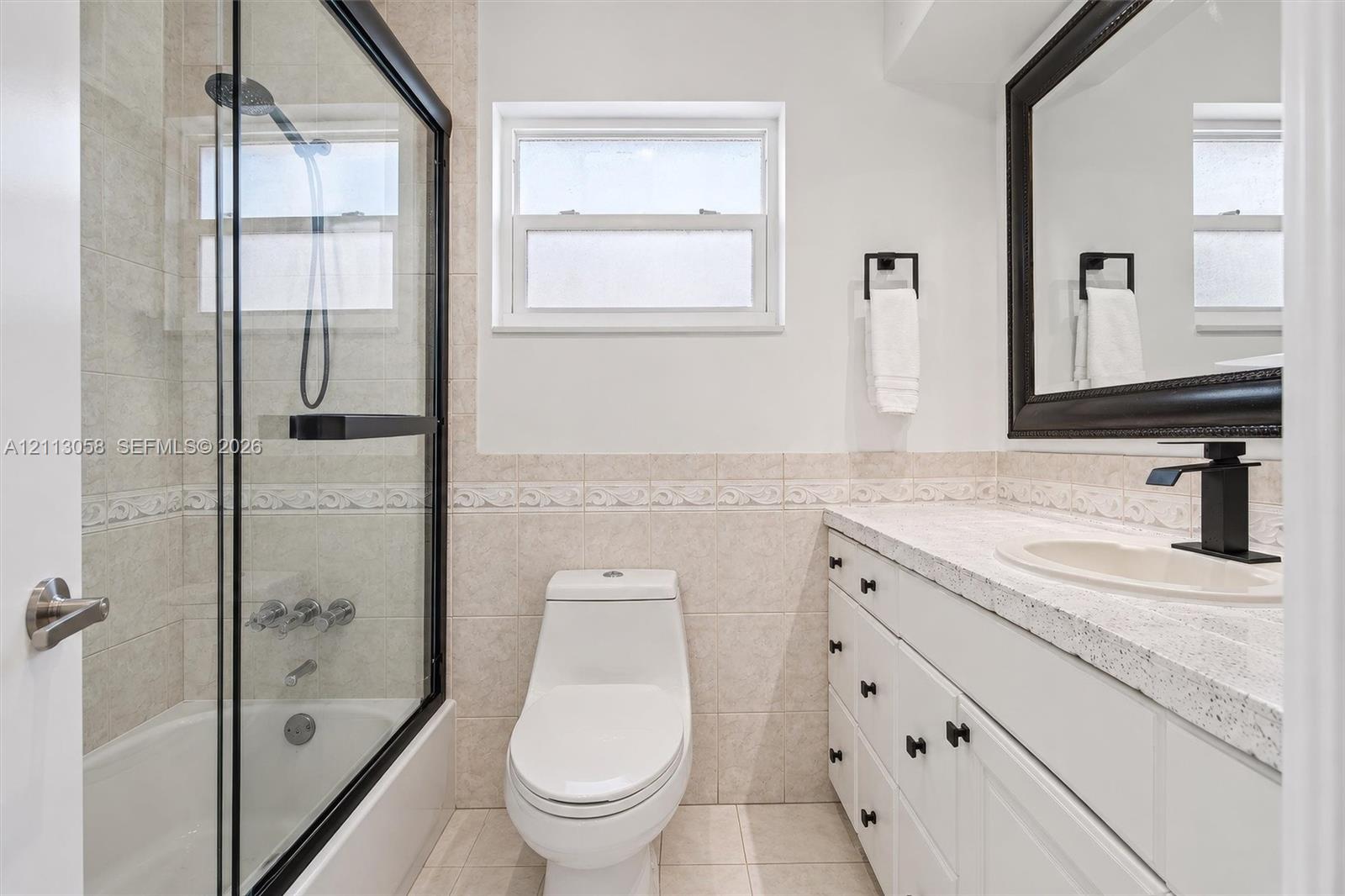 Bathroom featuring a shower-tub combination with a black-framed glass enclosure, vanity with speckled countertop and integrated basin, matte black faucet hardware, and neutral tile wall accents