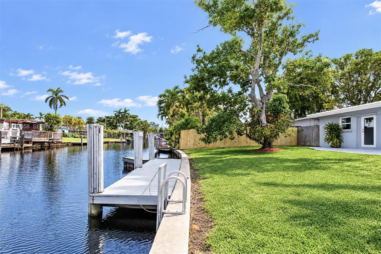 Waterfront backyard featuring a private dock, concrete seawall, manicured lawn, and mature trees