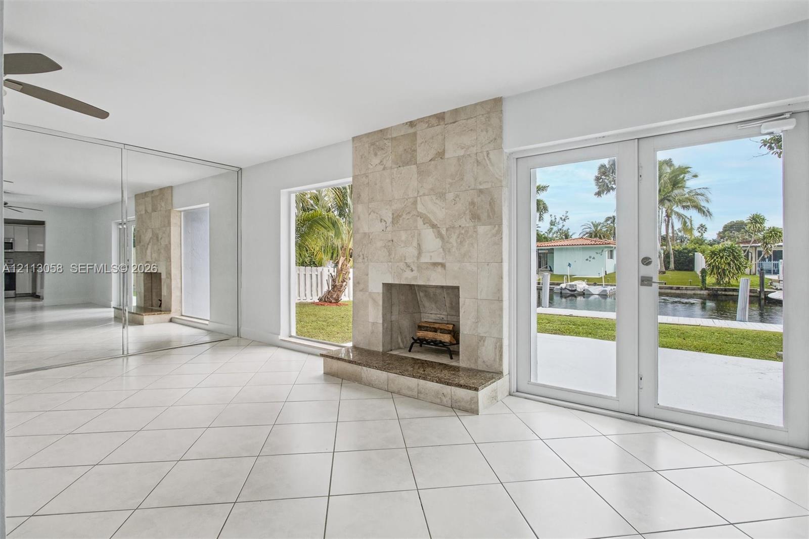 Neutral tile flooring, floor-to-ceiling mirrored wall panels, stone-tiled fireplace with a raised hearth, and double glass doors opening to a waterfront exterior
