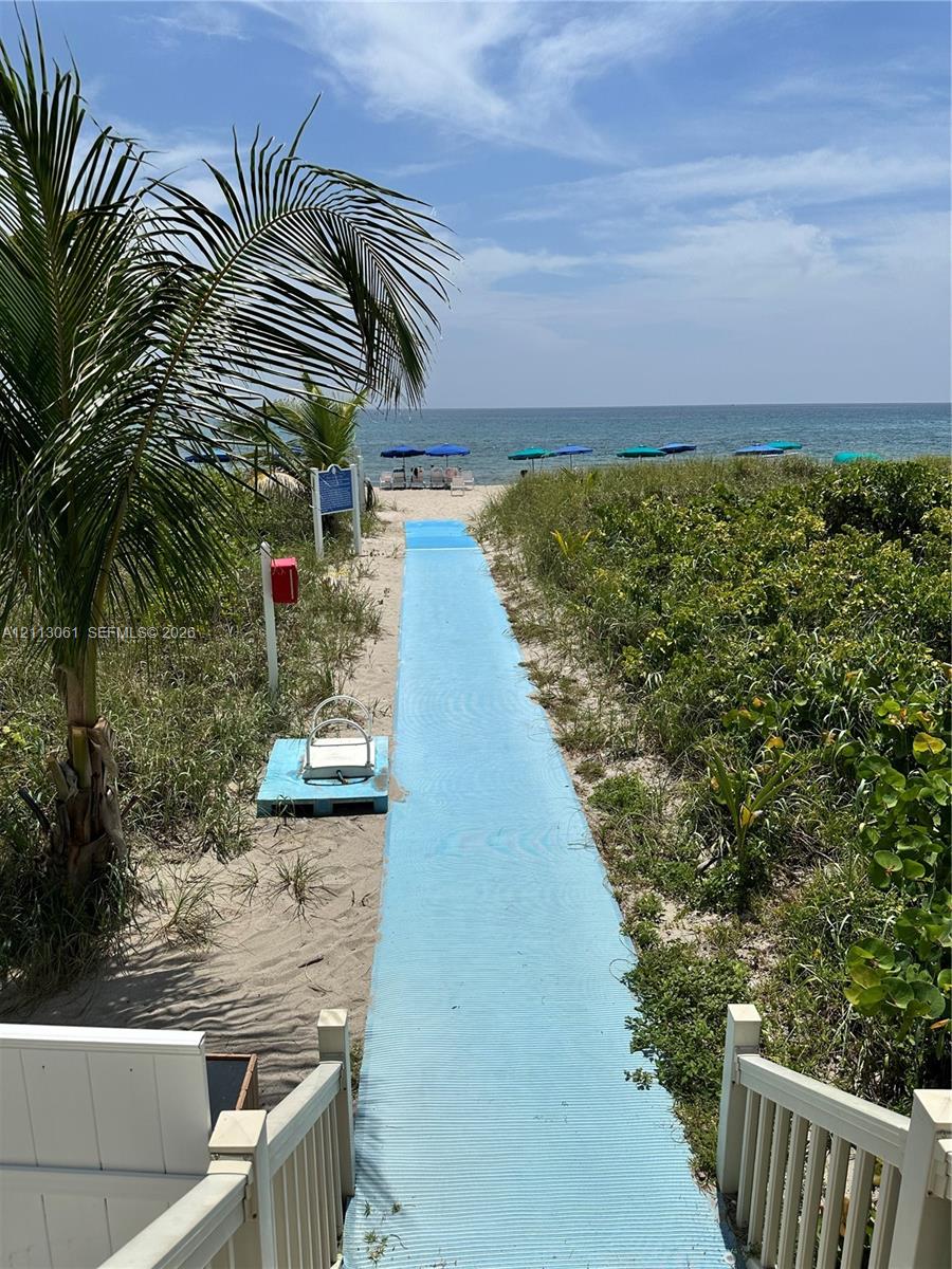 Direct beach access featuring a paved walkway, sandy shoreline, mature coastal vegetation, and a wooden staircase