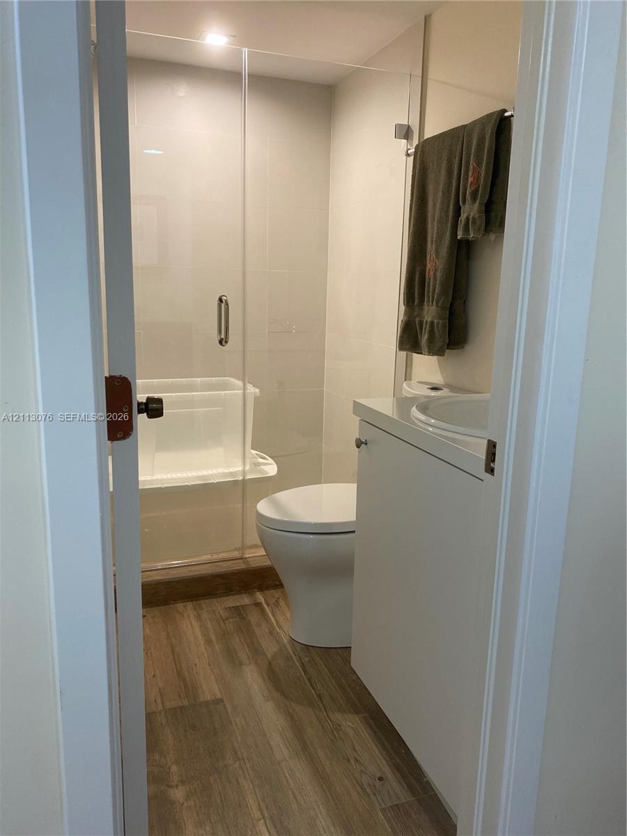 Bathroom featuring a walk-in shower with a glass enclosure, wood-finish flooring, a white vanity with an integrated sink, and a high-profile toilet