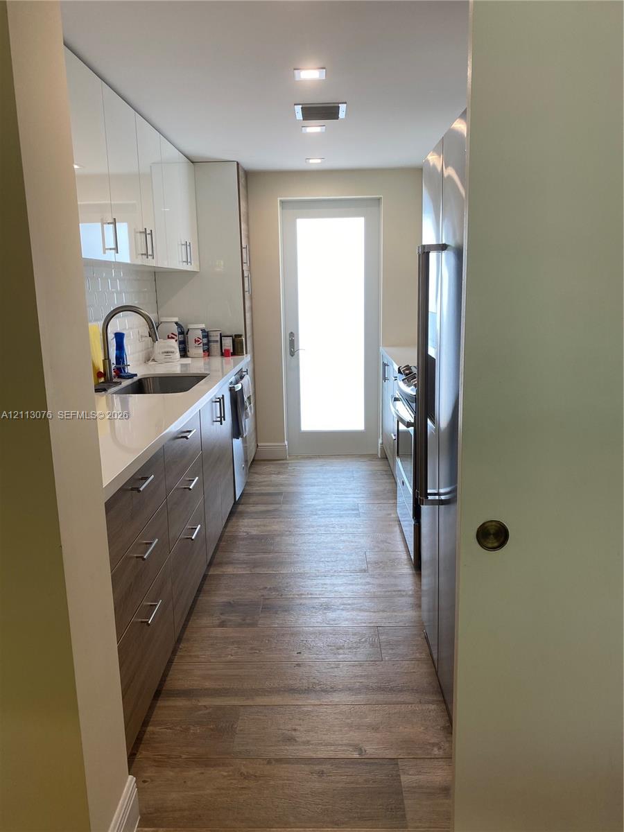 Galley kitchen featuring wood-finish cabinetry, white slab countertops, stainless steel appliances, wood-finish flooring, and a glass-paneled exterior door