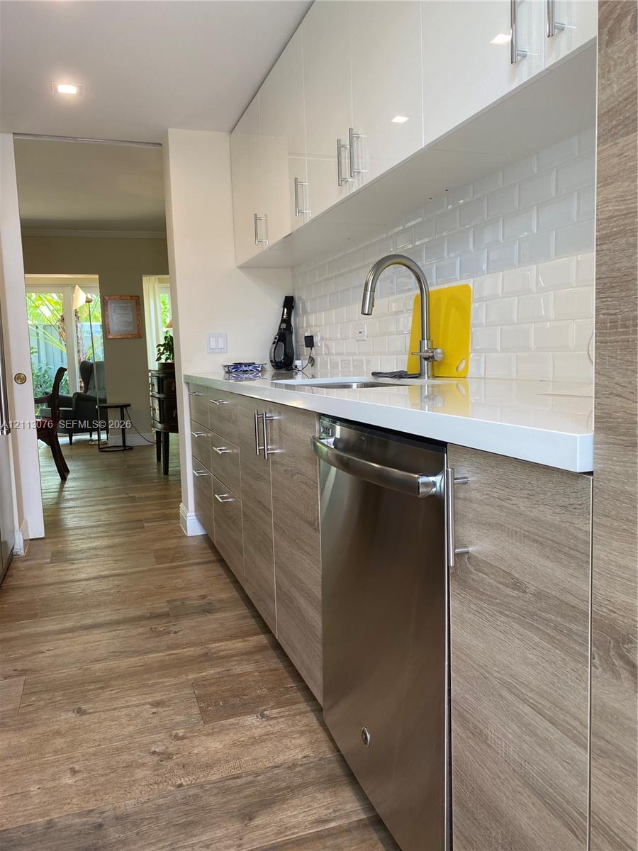 Kitchen featuring high-gloss white upper cabinetry, wood-finish lower cabinetry, white solid-surface countertops, white subway tile backsplash, and stainless steel dishwasher