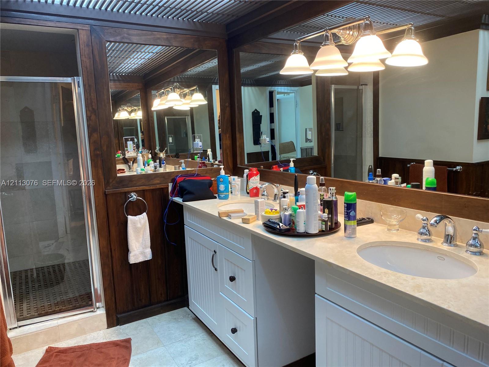 Dual-sink vanity with white cabinetry, stone-finish countertops, wall-to-wall mirrors with wood-trim, a glass-enclosed shower, and multi-light vanity fixtures